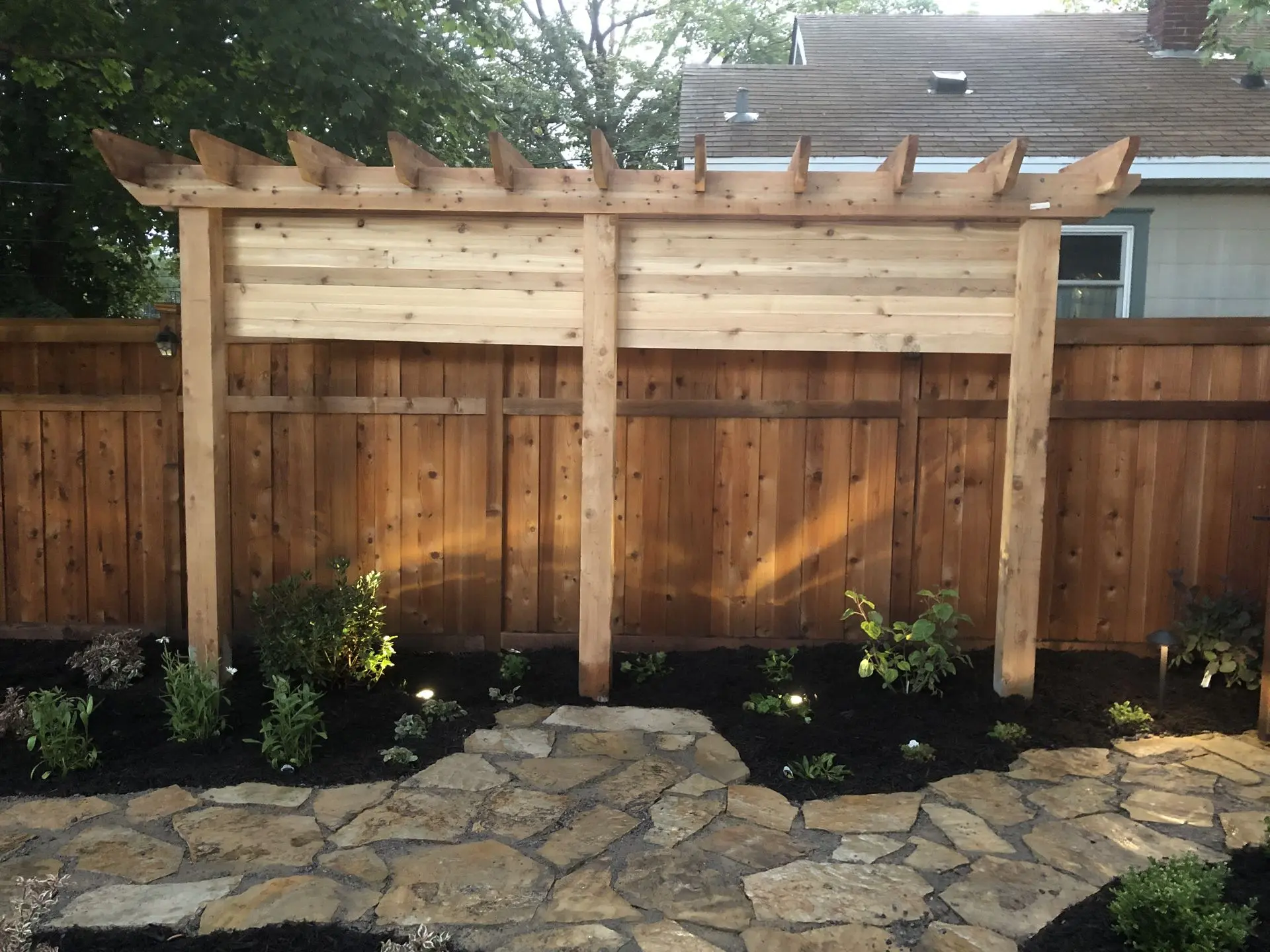 A custom built trellis style privacy fence, nestled near a natural stone paver walkway, creating a warm and private outdoor living area.