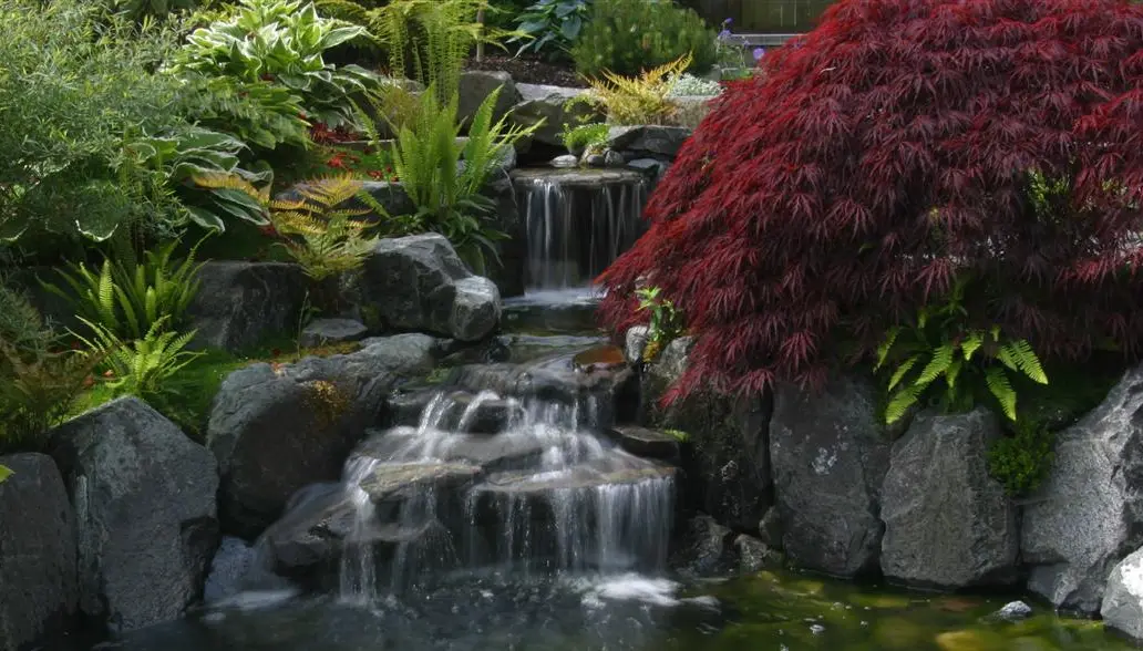 A rustic rock waterfall flowing into a stone-lined pond, adding a dynamic and natural element to the garden landscape.